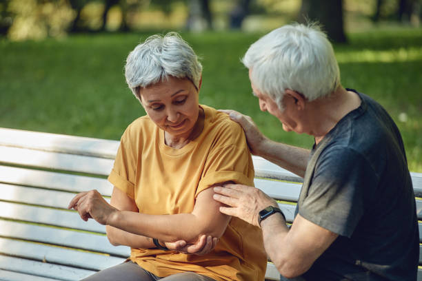 During morning sportive stroll or making exercises in park, old 60s woman got injured her shoulder or elbow gripping arm sit in bench with caring disappointed husband. Traumas of older people concept